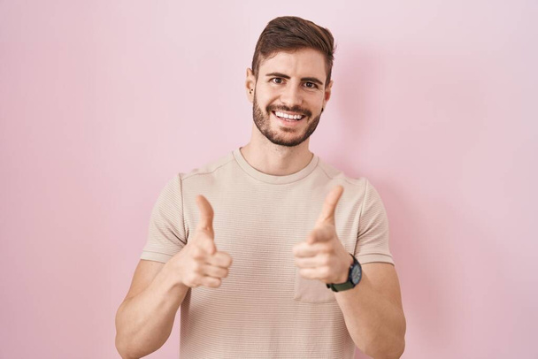 Hispánec s plnovousem stojící nad růžovým pozadím ukazující prsty na kameru se šťastnou a zábavnou tváří. dobrá energie a vibrace.  - Fotografie, Obrázek