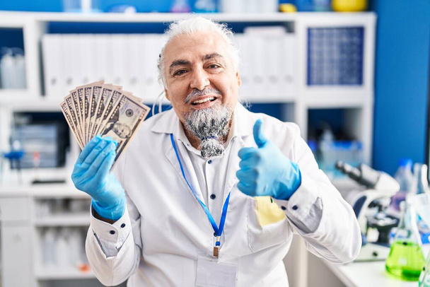 Muž středního věku s šedými vlasy pracuje v laboratoři vědec drží peníze s úsměvem šťastný a pozitivní, palec nahoru dělá vynikající a schvalovací znamení  - Fotografie, Obrázek
