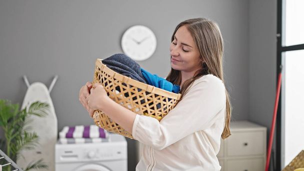 Joven hermosa mujer hispana sonriendo confiada sosteniendo canasta de mimbre con ropa en la lavandería - Foto, Imagen