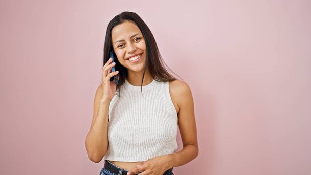 Junge schöne hispanische Frau lächelt selbstbewusst und spricht auf dem Smartphone vor isoliertem rosa Hintergrund - Foto, Bild
