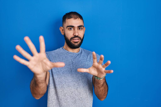 Middle east man with beard standing over blue background afraid and terrified with fear expression stop gesture with hands, shouting in shock. panic concept.  - Foto, afbeelding
