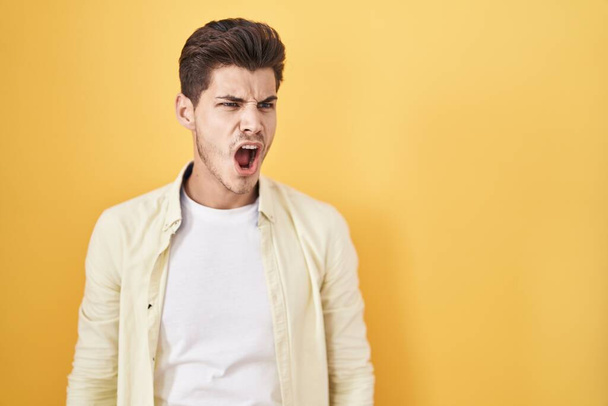 Young hispanic man standing over yellow background angry and mad screaming frustrated and furious, shouting with anger. rage and aggressive concept.  - 写真・画像