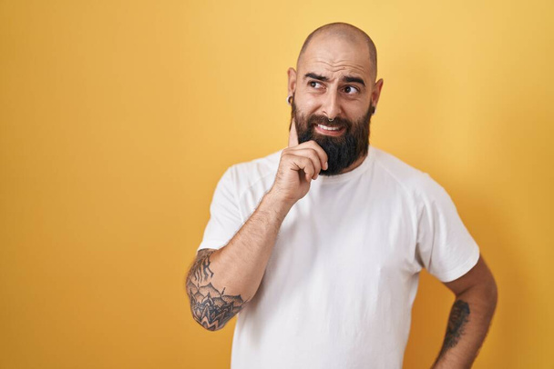 Mladý hispánec s vousy a tetováním stojí nad žlutým pozadím a přemýšlí o otázce, znepokojen a nervózní s rukou na bradě  - Fotografie, Obrázek