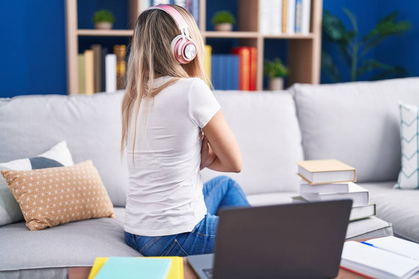 Jeune femme blonde étudiant à l'aide d'un ordinateur portable à la maison debout vers l'arrière regardant loin avec les bras croisés  - Photo, image