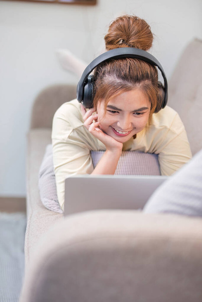 Młoda piękna azjatycka kobieta relaksująca się i słuchająca muzyki za pomocą słuchawek, leży na kanapie. Młoda kobieta korzystających w domu za pomocą laptopa..... - Zdjęcie, obraz