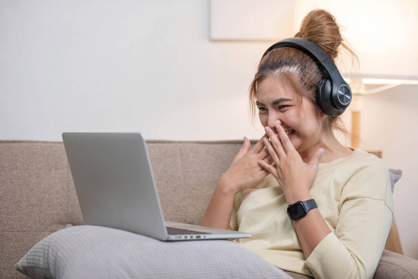 Jeune belle femme asiatique se détendre et écouter de la musique à l'aide d'écouteurs, elle est allongée dans un canapé. Jeune femme appréciant à la maison en utilisant un ordinateur portable..... - Photo, image