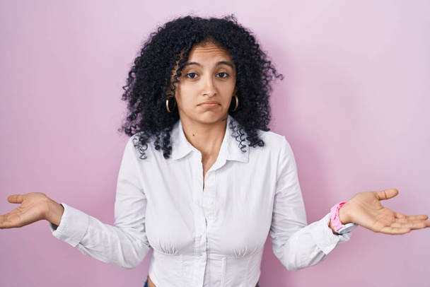 Hispanic woman with curly hair standing over pink background clueless and confused expression with arms and hands raised. doubt concept.  - Fotoğraf, Görsel