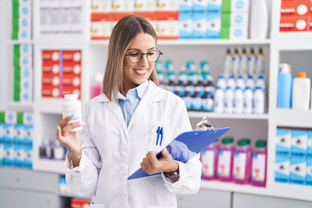 Jonge mooie Spaanse vrouw apotheker holding pillen fles lezen document in de apotheek - Foto, afbeelding