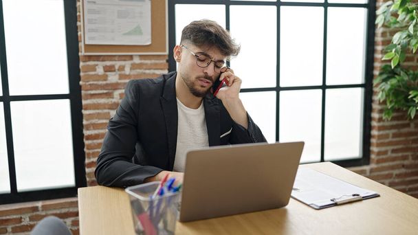 Молодой арабский бизнесмен с помощью ноутбука разговаривает на смартфоне в офисе - Фото, изображение