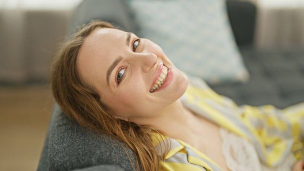 Mujer rubia joven relajada en el sofá sonriendo en casa - Foto, Imagen