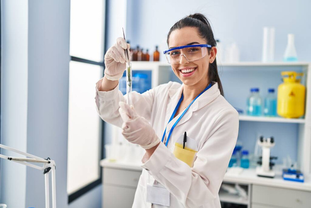 Young beautiful hispanic woman scientist holding plant with tweezer at laboratory - Photo, Image