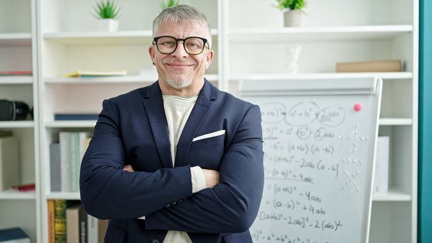 Moyen âge homme aux cheveux gris enseignant souriant confiant debout avec les bras croisés geste par tableau blanc à l'université salle de classe - Photo, image