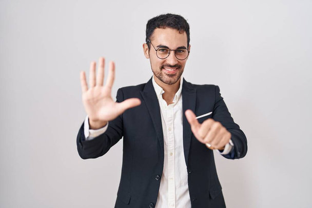 Bonito homem hispânico de negócios em pé sobre fundo branco mostrando e apontando para cima com os dedos número seis, sorrindo confiante e feliz.  - Foto, Imagem