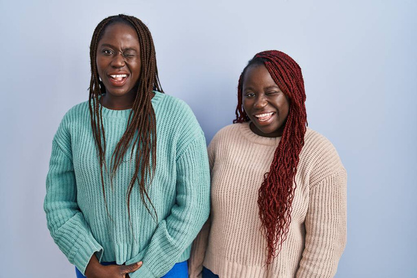 İki Afrikalı kadın mavi arka planda dikiliyor ve kameraya seksi bir ifadeyle bakıyor, neşeli ve mutlu bir yüzle..  - Fotoğraf, Görsel