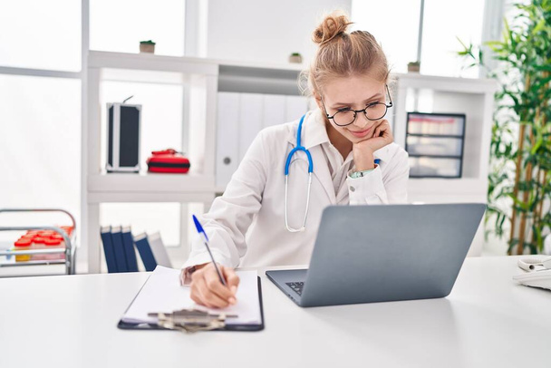 Mujer joven rubia médico usando portátil escribir informe médico en la clínica - Foto, imagen