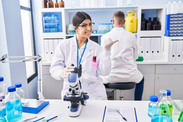 Jovem mulher trabalhando no laboratório cientista apontando o polegar para o lado sorrindo feliz com a boca aberta  - Foto, Imagem