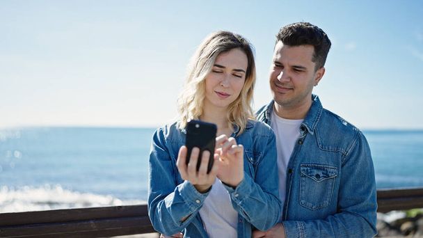 Мужская и женская пара стоят вместе, используя смартфон на берегу моря - Фото, изображение