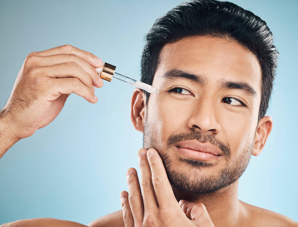 Face, beauty and antiaging oil with a man in studio on a blue background for skincare or grooming. Luxury, serum or wellness with a handsome male person in the bathroom to apply a product to his face. - 写真・画像