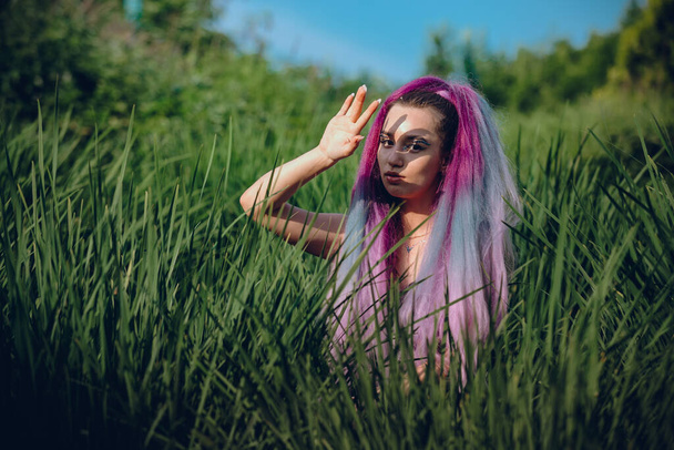 Uzun menekşe renkli saçları olan genç bir kadın, çayırda uzun yeşil çimlerle oturuyor, elini yüzüne kaldırıyor, güneş ışığını engelliyor, yüzünde bir gölge yaratıyor. - Fotoğraf, Görsel
