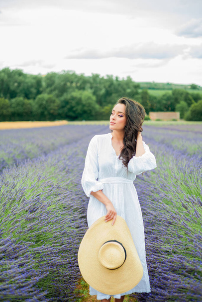 Lavender field. Beautiful dark-haired curly woman in white simple dress in field of purple flowers. - Photo, Image