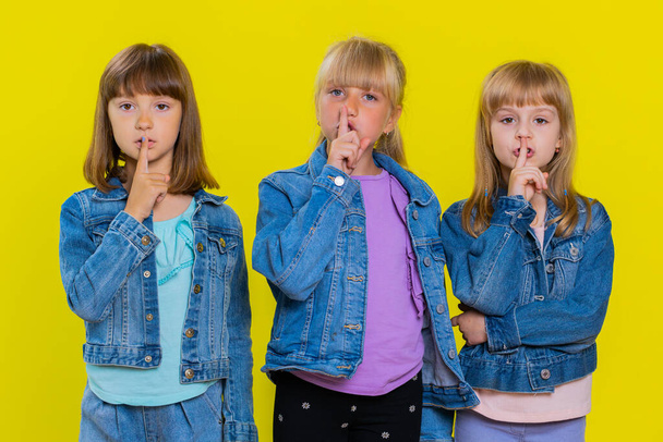 Shh be quiet please. Teenage cute girls presses index finger to lips makes silence gesture sign do not tells secret. Little children sisters. Three siblings kids isolated on studio yellow background - Photo, image
