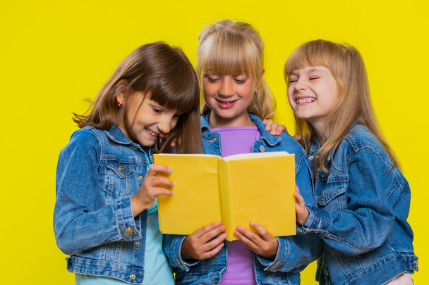 Teenage girls reading funny interesting fairytale story book, leisure hobby, knowledge wisdom, education, learning, study, wow. Little children sisters. Three cute siblings kids on yellow background - Fotoğraf, Görsel