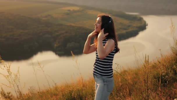 Ein junges Mädchen vor dem Hintergrund der Natur des Flusses und der Berge genießt bei Sonnenuntergang Musik mit Kopfhörern, singt mit und tanzt fröhlich und amüsant in die Kamera - Filmmaterial, Video