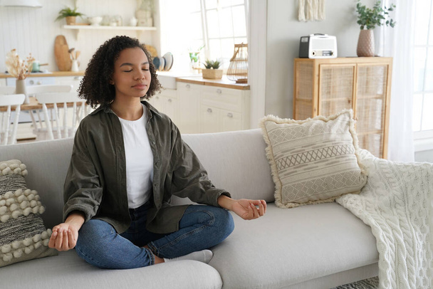 Serene mixed-race girl meditates at home, in lotus pose with mudra gesture on the sofa. Afro-haired, calm, practicing yoga. Healthy lifestyle, meditation. - Photo, Image