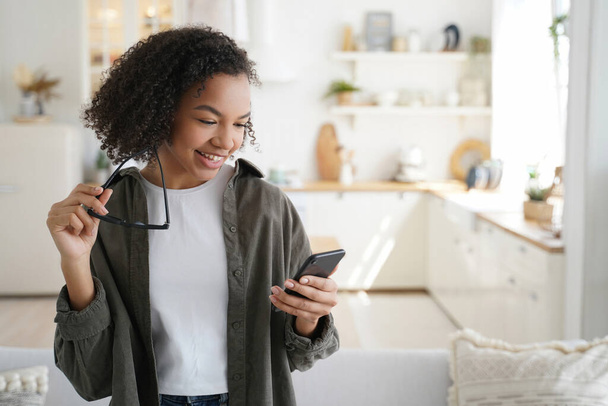 Smiling mixed-race girl with afro hairstyle using smartphone, shopping online, and chatting on social networks. Modern teen, reading good news at home. - Φωτογραφία, εικόνα