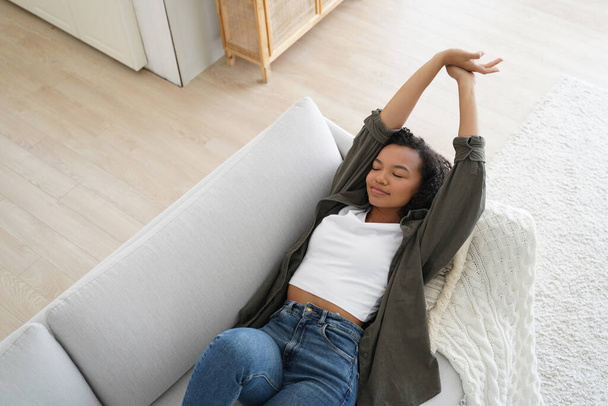 Heiteres afroamerikanisches Mädchen, das zu Hause auf einem bequemen Sofa schläft und an einem faulen Wochenende eine entspannende Pause genießt. Entspannte junge Frau streckt Arme aus, ruht sich aus und liegt in einem gemütlichen Wohnzimmer. - Foto, Bild