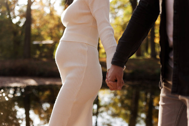 Пара, чоловік і вагітна дружина тримаються за руки і ходять в осінній парку на заході сонця. Щаслива родина, яка любить вагітність. Поняття Дня матері і жіночого дня - Фото, зображення
