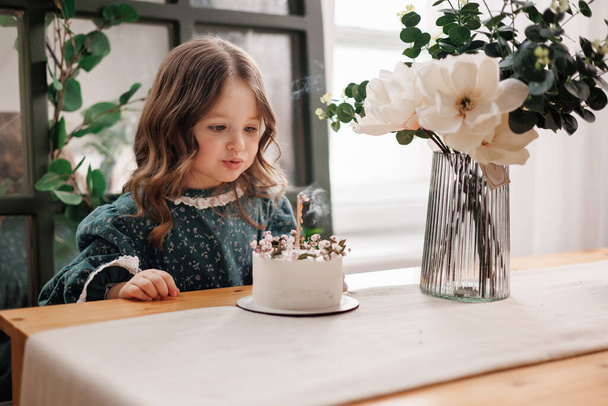 Festa de aniversário infantil. Menina adorável senta-se à mesa com flores decoradas bolo de aniversário e queimar vela, faz o desejo. Criança em vestido festivo no quarto com balões, buquê de flores no vaso. - Foto, Imagem