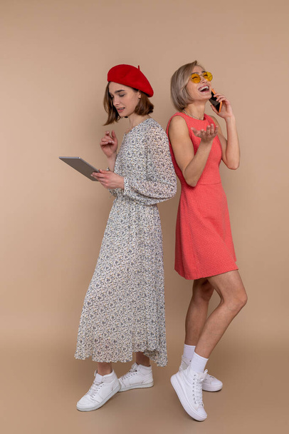 Two women friends wearing elegantdresses standing together isolated over beige background using digital tablet and mobile phone. - Фото, изображение