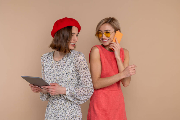 Zarif elbiseler giyen iki kadın arkadaş dijital tablet ve cep telefonu kullanarak bej arkaplan üzerinde izole bir şekilde duruyorlar.. - Fotoğraf, Görsel