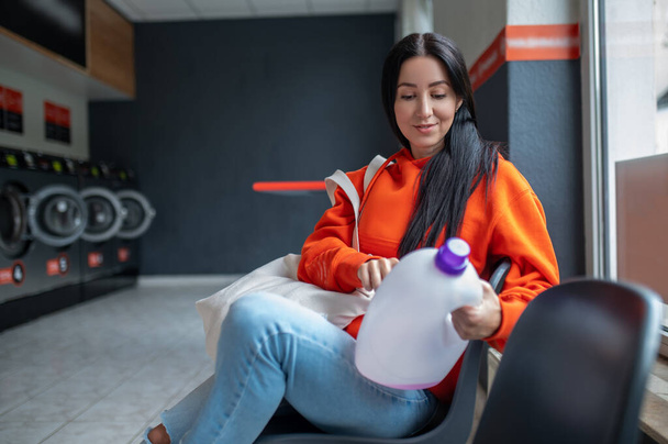 Young woman wearing orange hoodie with bag reading how to use washingdetergent in public laundry. - Foto, imagen