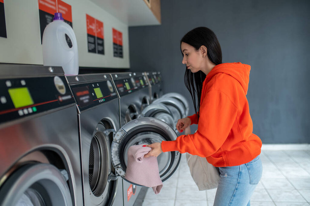 Brunette woman inserting dirty clothes into the washing machine in the laundry room. - Photo, image