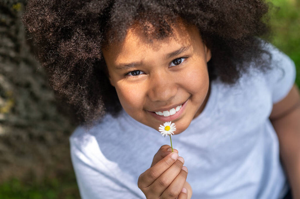 Happy smiling little girl child outdoors showing little chamomile flower while resting in sunny summer park. - Foto, immagini