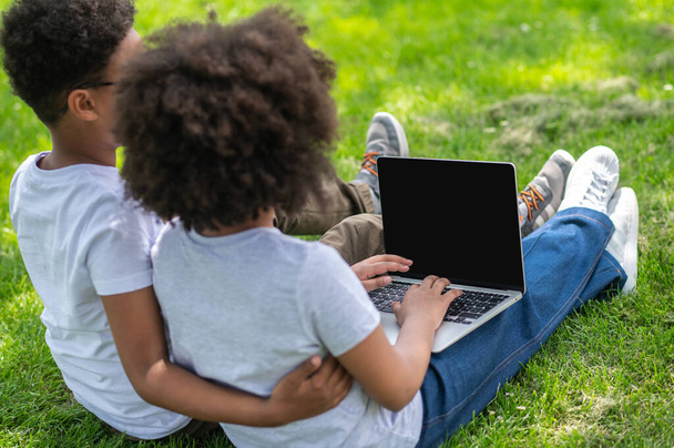 Little children friends girl and boy with laptop sitting on grass watching movie browsing internet, blank screen. - Fotó, kép
