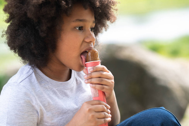 Little girl with curly hair wearing white t shirt eating delicious chocolate ice cream in garden and enjoys on hot day. - Fotó, kép