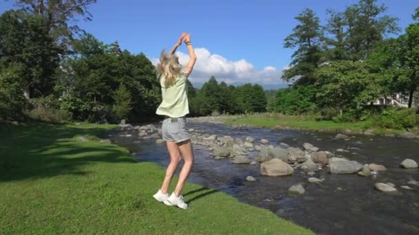 ショートパンツとTシャツの若い美しい女性は,山の川で喜びと緑と山を見下ろすためにジャンプします. 旅行のコンセプト,週末は自然に出かけています. 都市の後の荒野を楽しむ. - 映像、動画
