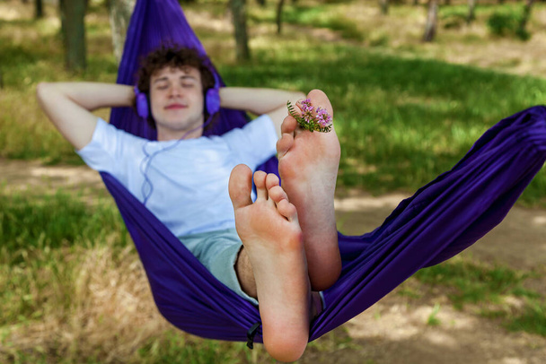 A young guy lying in a purple hammock listens to music on headphones while enjoying nature. - 写真・画像