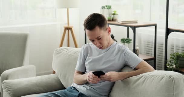 Homem caucasiano alegre está sentado em um sofá usando uma camiseta casual. Ele está envolvido com o telefone, conversando com alguém ou navegando conteúdo on-line, e ele faz isso com um sorriso brilhante em seu rosto. - Filmagem, Vídeo