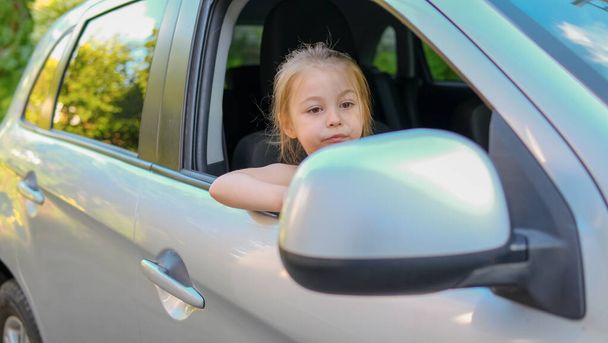 Szczęśliwa dziewczynka wyglądająca przez okno samochodu uśmiechnięta do kamery pozowanie jazdy z rodzicami w samochodzie po drugiej stronie miasta. Auto podróży i przygody drogowej Concept. Wysokiej jakości zdjęcie - Zdjęcie, obraz