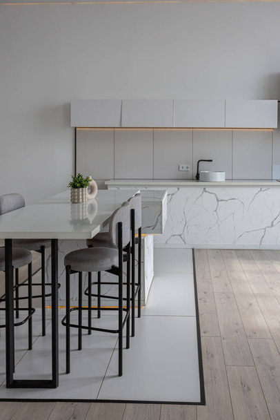 kitchen and dining island with high bar stools in a minimalistic modern luxury design of an expensive house on a sunny day. white walls with plaster, parquet, decorative lighting and no one inside - Φωτογραφία, εικόνα