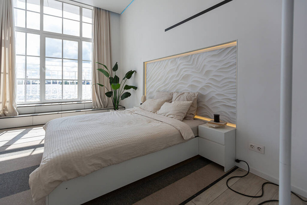 bedroom area in a minimalistic modern luxury design of an expensive house on a sunny day. decorative panel in the headboard, white walls with plaster, parquet, decorative lighting and no one - Foto, afbeelding