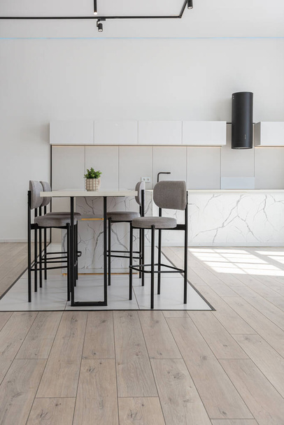 kitchen and dining island with high bar stools in a minimalistic modern luxury design of an expensive house on a sunny day. white walls with plaster, parquet, decorative lighting and no one inside - Photo, Image
