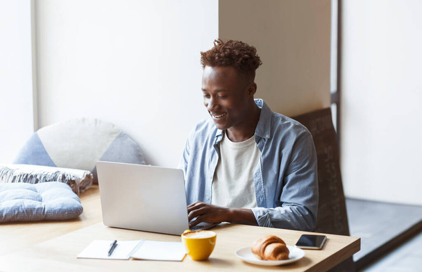 Online arbeiten, Fernjob-Konzept. Cooler positiver Millennial Afroamerikaner in lässigem Outfit sitzt im Café, trinkt Kaffee, isst Croissant, benutzt Laptop, macht freiberufliche Arbeit, kopiert Raum - Foto, Bild