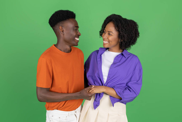 Cheerful millennial african american man and lady in casual hugging, holding hands, enjoy romantic date, isolated on green studio background. Relationship, love and walk, ad and offer - Foto, immagini