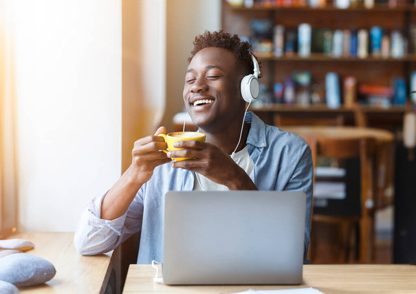 陽気なアフリカ系アメリカ人の百万人の男でカジュアルに彼の朝のコーヒーを楽しみながら、音楽やカフェでお気に入りのポッドキャストを聞いて、黄色のマグカップと笑い、空白のスペース、太陽のフレアを保持 - 写真・画像