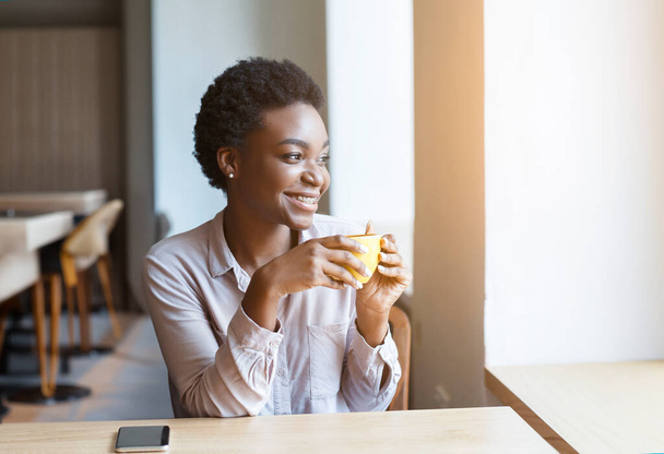 Alegre bela milenar senhora afro-americana em roupa casual inteligente relaxante com xícara de café delicioso aromático no café acolhedor, olhando através da janela e sorrindo, espaço de cópia, brilho do sol - Foto, Imagem